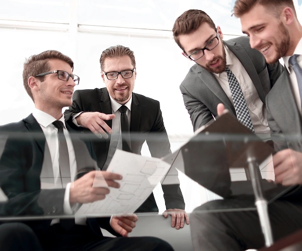 Czterech mężczyzn w garniturach współpracuje w biurze, przeglądając dokumenty i tablet. Wyglądają na zaangażowanych i skupionych, emanując poczuciem pracy zespołowej i profesjonalizmem.